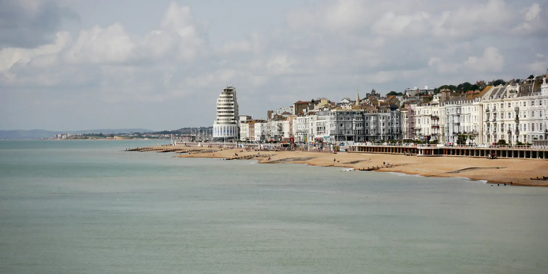 Coastal cityscape with a sandy beach, a modern cylindrical building, and Victorian townhouses under a partly cloudy sky.