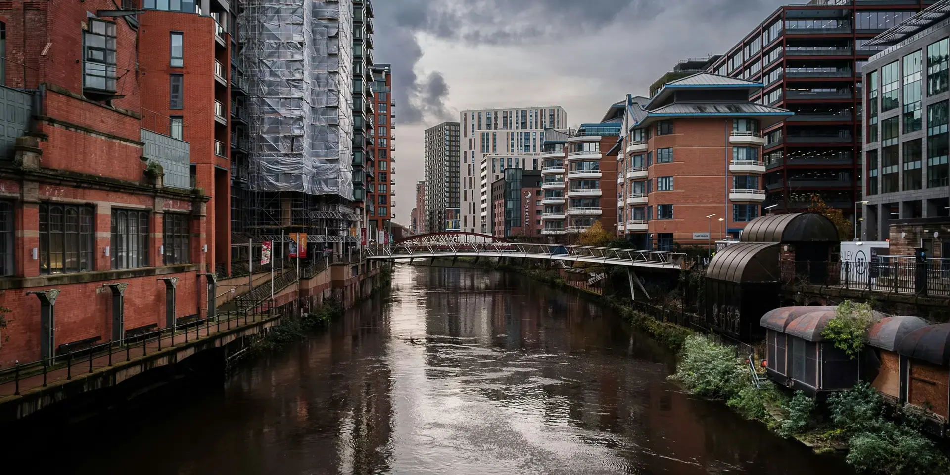 Modern buildings line the river in Manchester’s city center with a pedestrian bridge connecting both sides under a cloudy sky