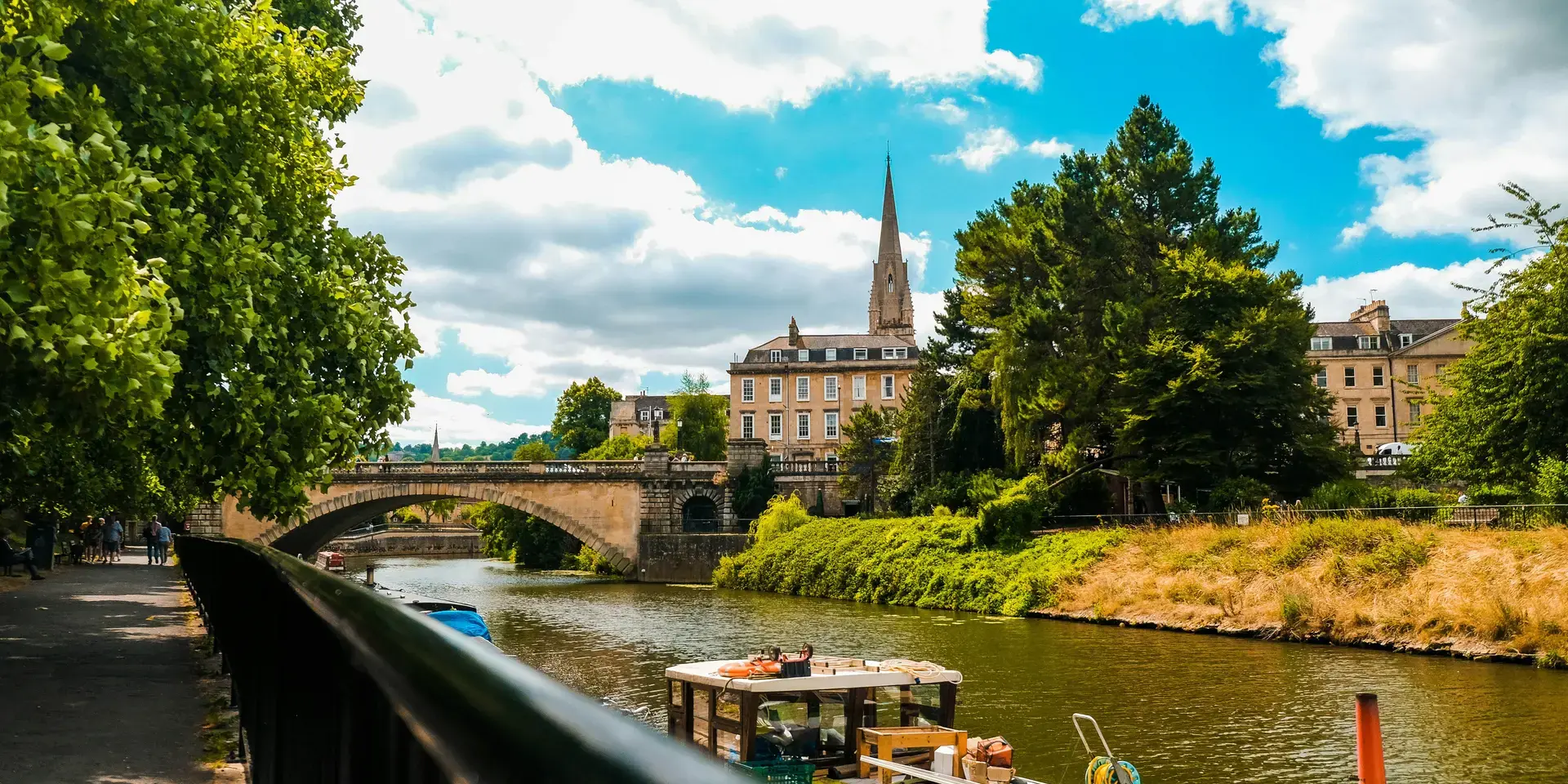Riverside view with lush trees, a stone bridge, and a church spire rising behind historic buildings under a bright blue sky