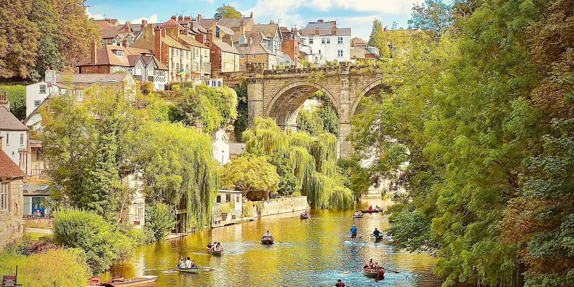 Historic stone bridge over a river surrounded by charming townhouses and lush trees, with people enjoying rowing boats