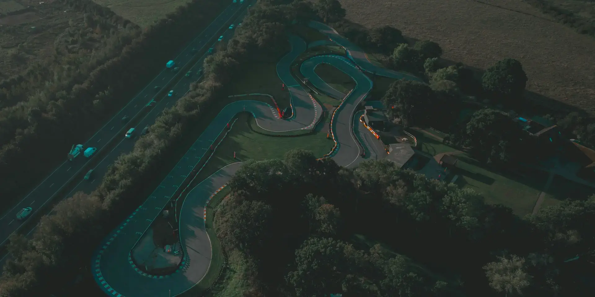 Aerial view of a winding karting track surrounded by greenery, next to a busy highway under a soft evening sky