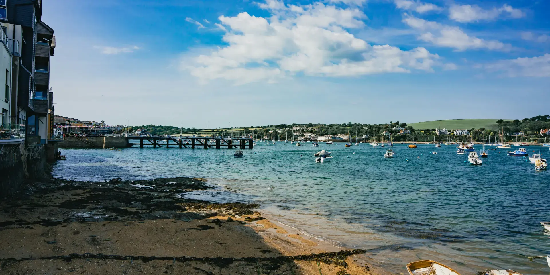 Small boats rest on a sandy shore by a harbor under a bright blue sky,with distant green hills and buildings across the water