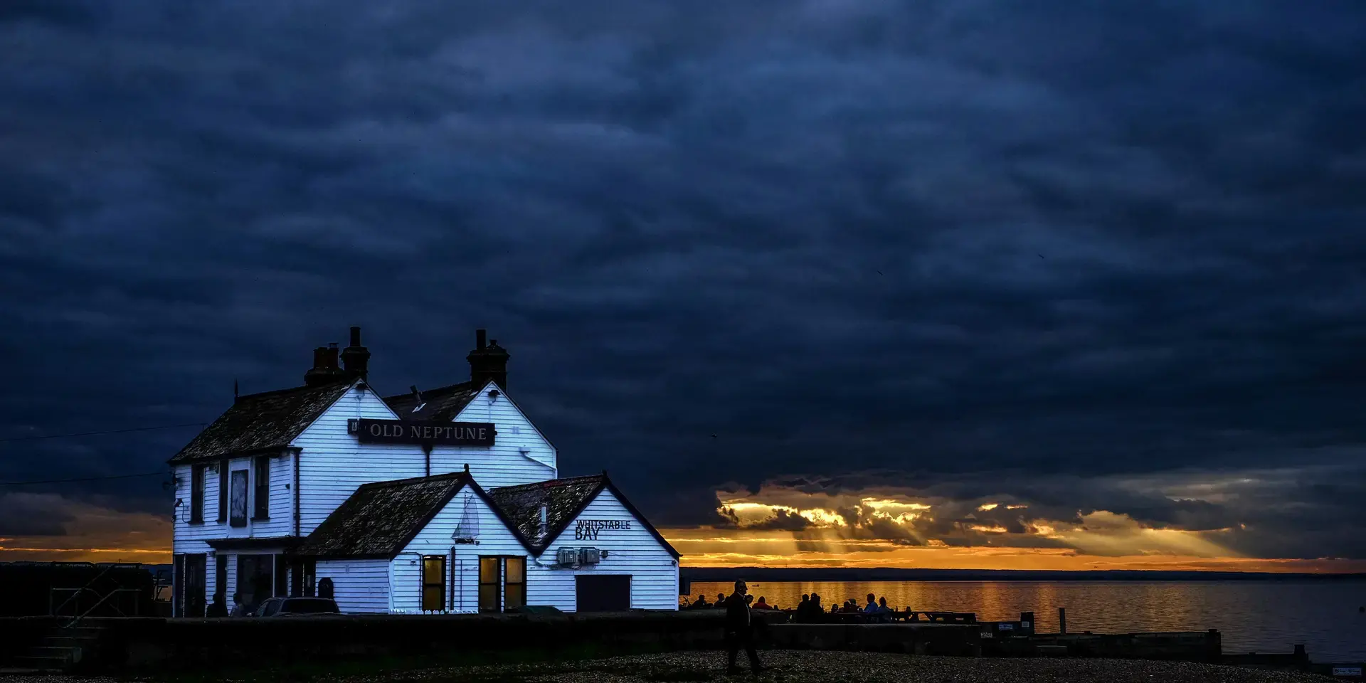 ‘Old Neptune’ pub at twilight, dramatic clouds above, golden sunset rays breaking over calm waters with silhouetted visitors.