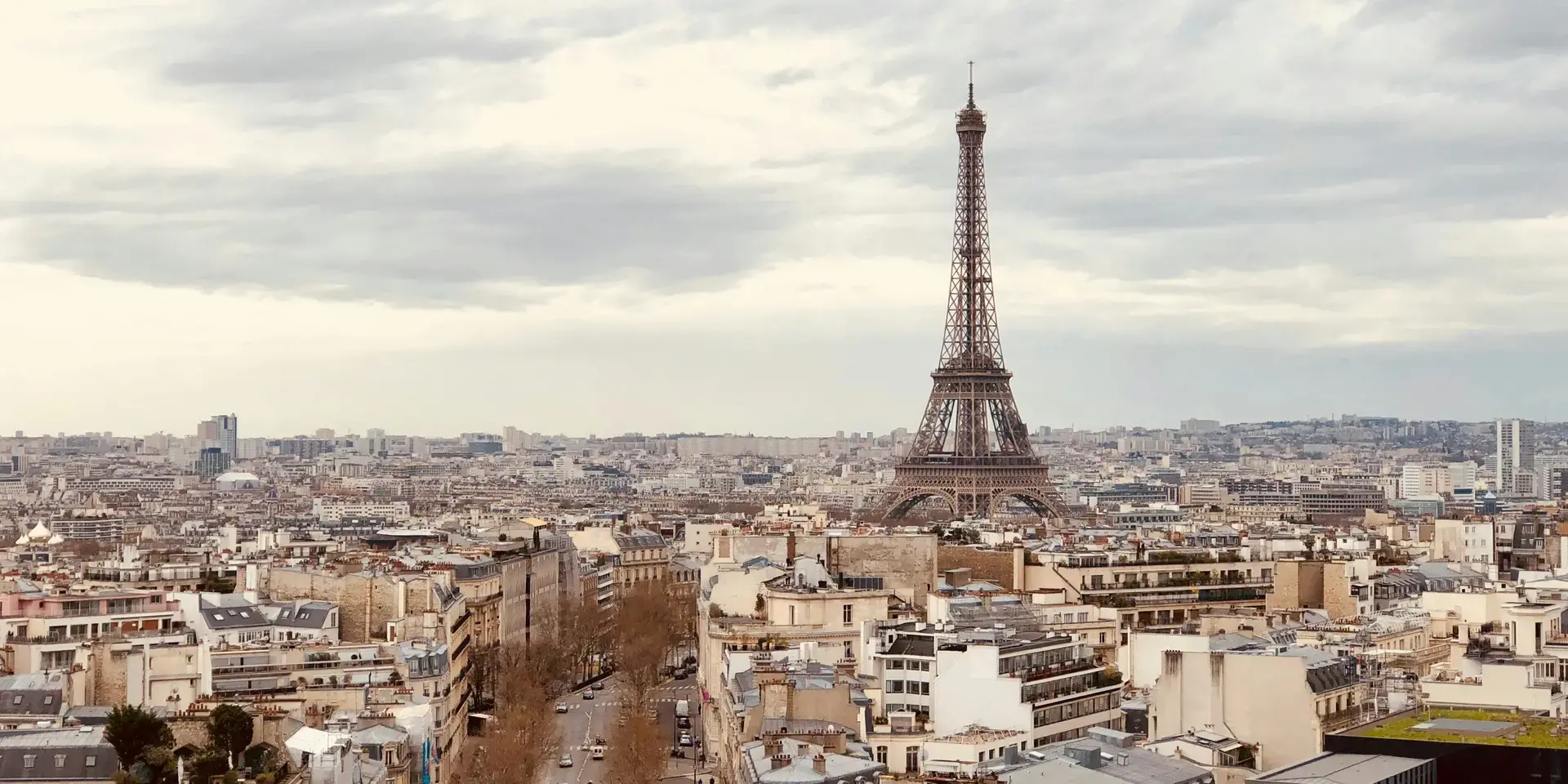 The Eiffel Tower rises over Paris under a cloudy sky, surrounded by historic rooftops and tree-lined avenues.