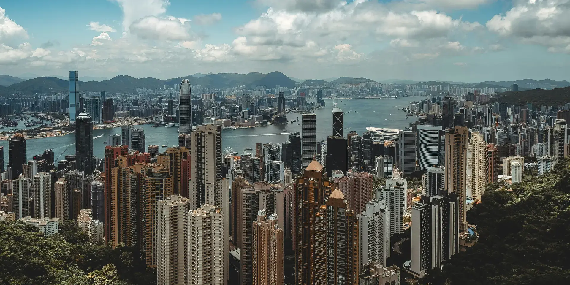 Skyline of Hong Kong with its towering skyscrapers, Victoria Harbour, and lush green hills under a bright, cloudy sky