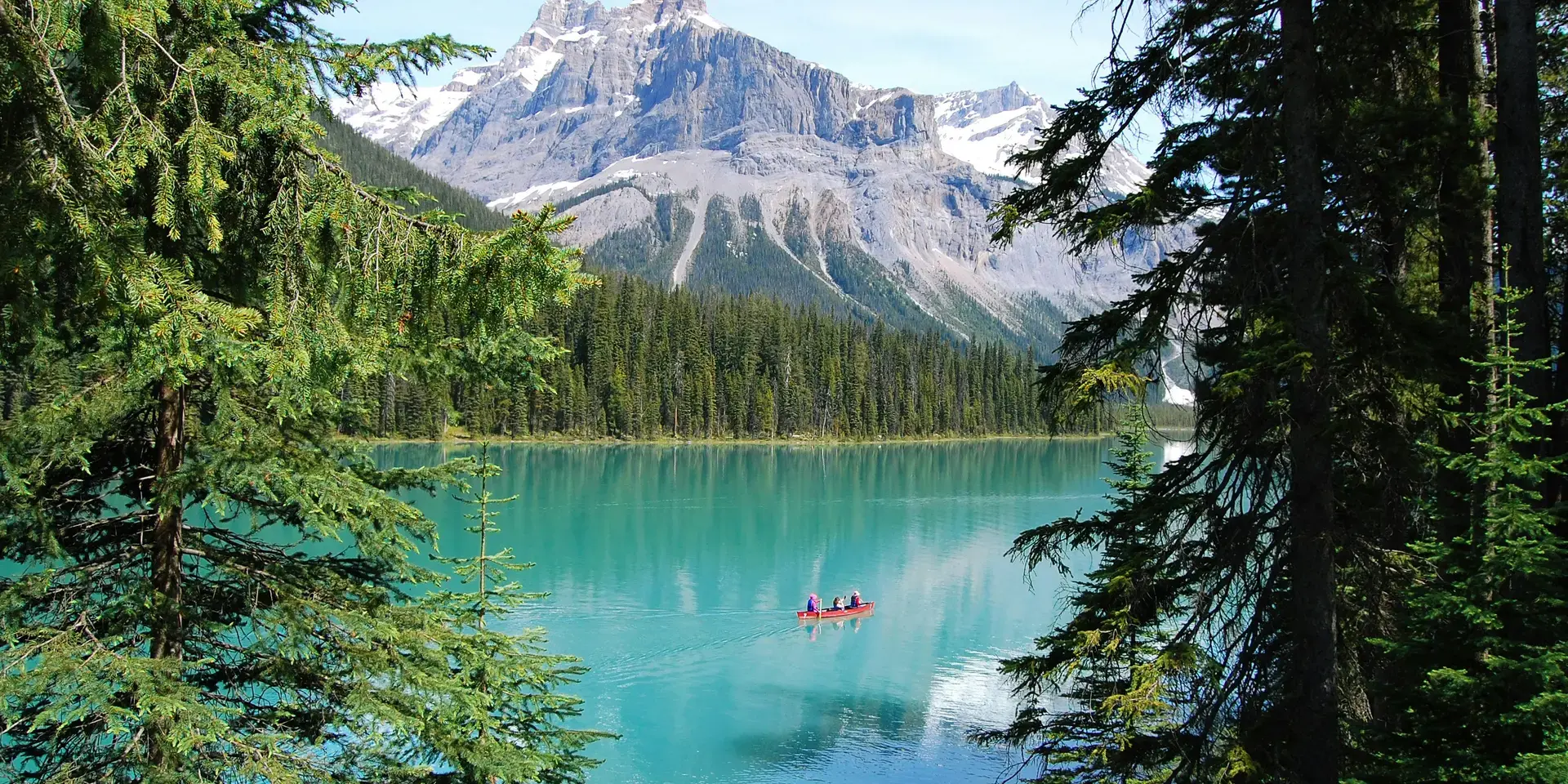 Turquoise lake with pine trees, snow-capped mountains, and a red canoe gliding peacefully on the water.