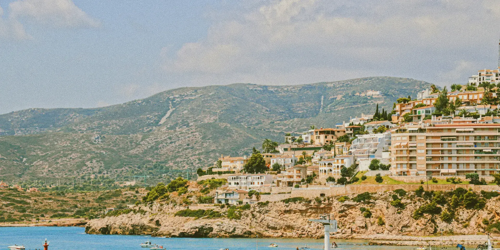 Scenic Mediterranean harbor with hillside homes, boats, and a red lighthouse under a bright, clear sky