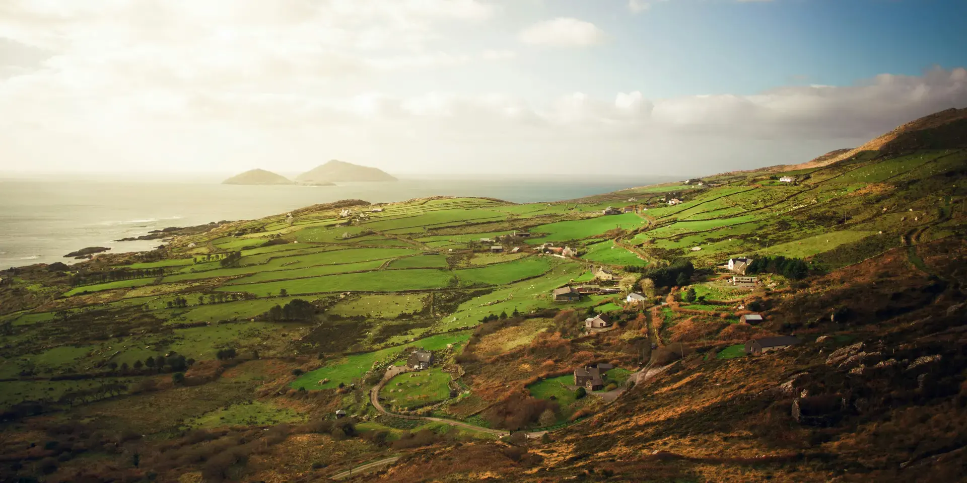 Rolling green Irish hills with scattered cottages overlooking the ocean, basking in soft sunlight under a partly cloudy sky.