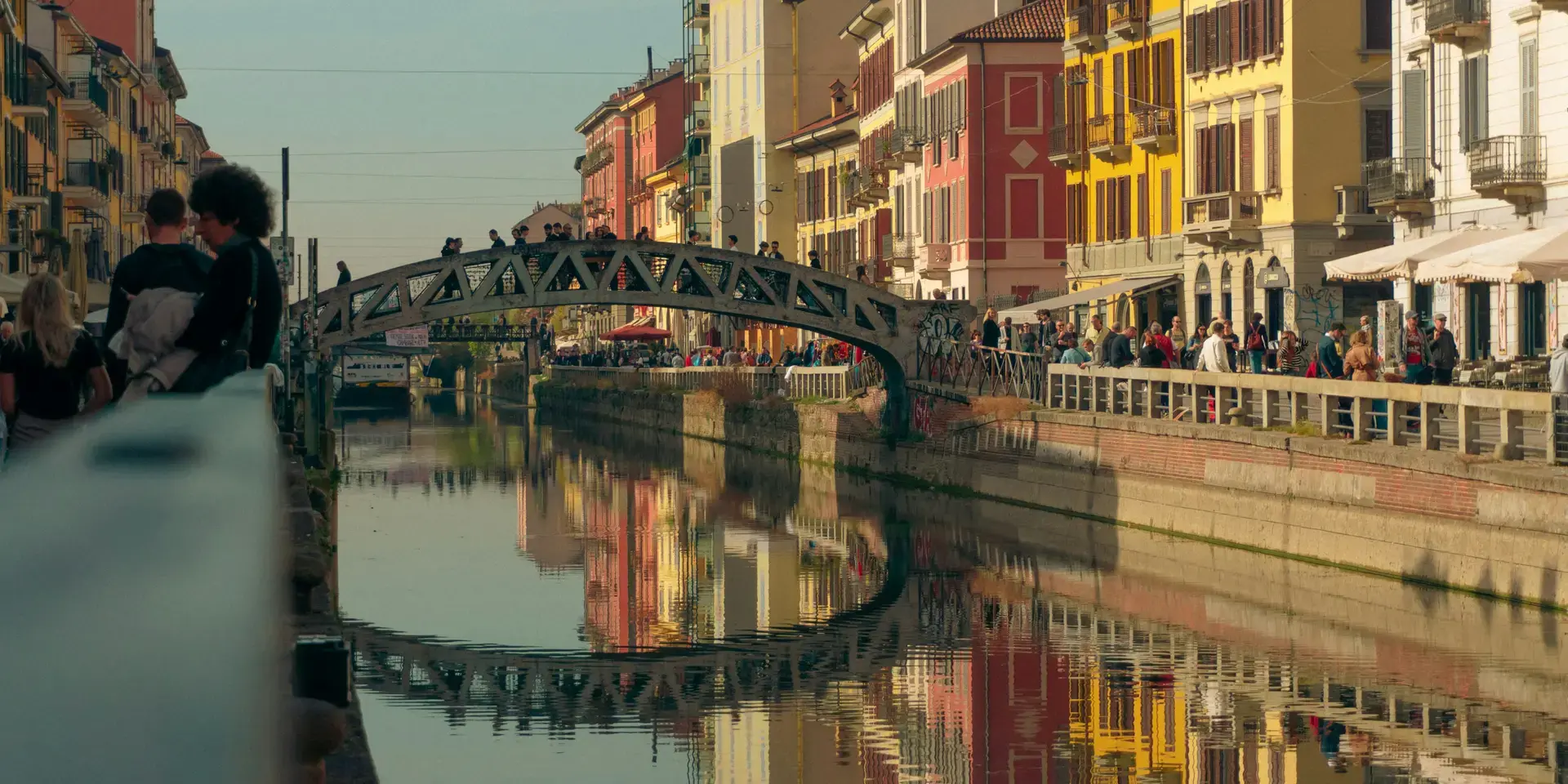 A lively Milan canal in Navigli, with colorful buildings reflecting in the water and people strolling along the promenade.