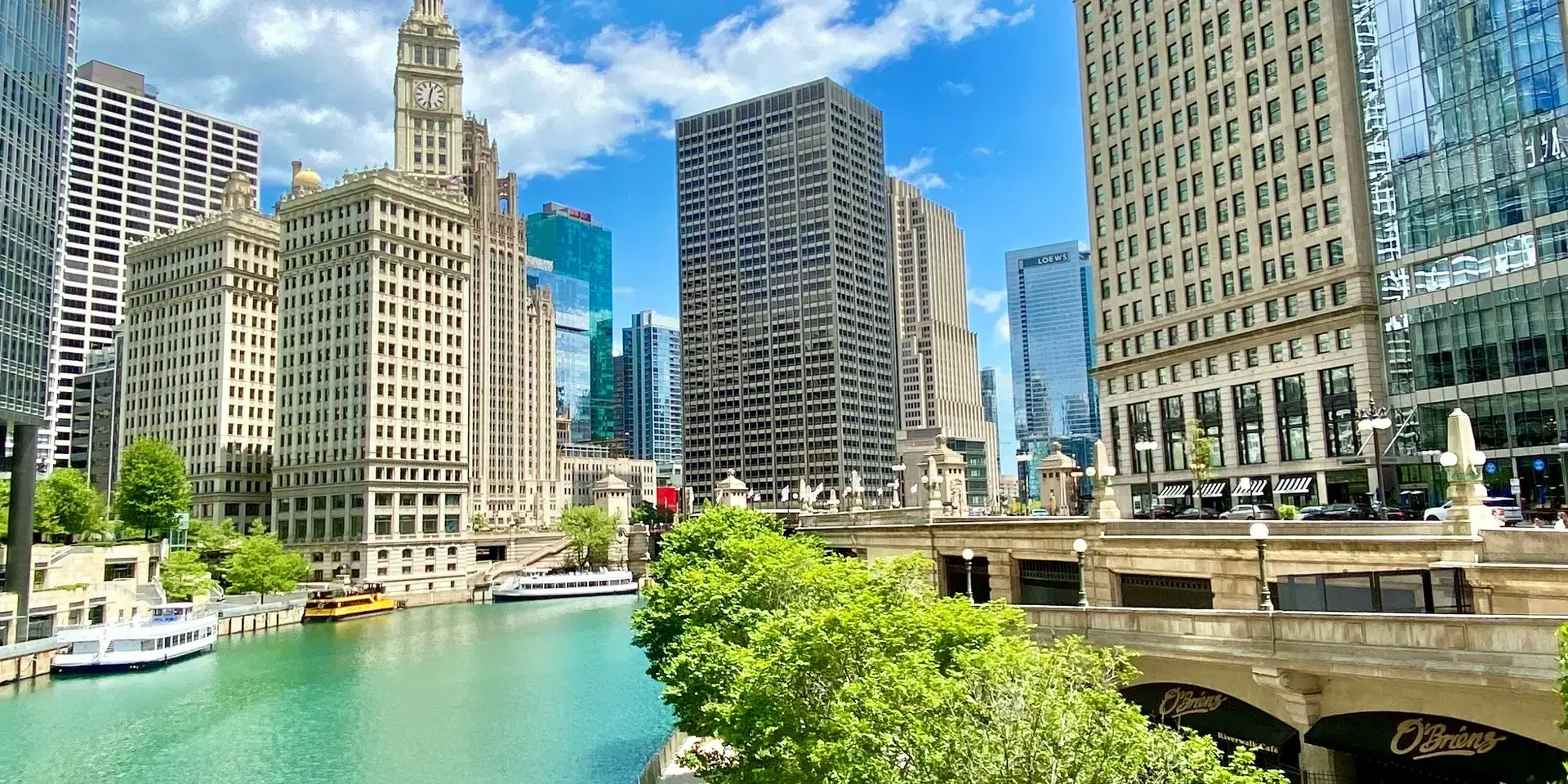 Chicago Riverwalk on a sunny day, with skyscrapers reflecting on the turquoise water and lush green trees lining the riverban