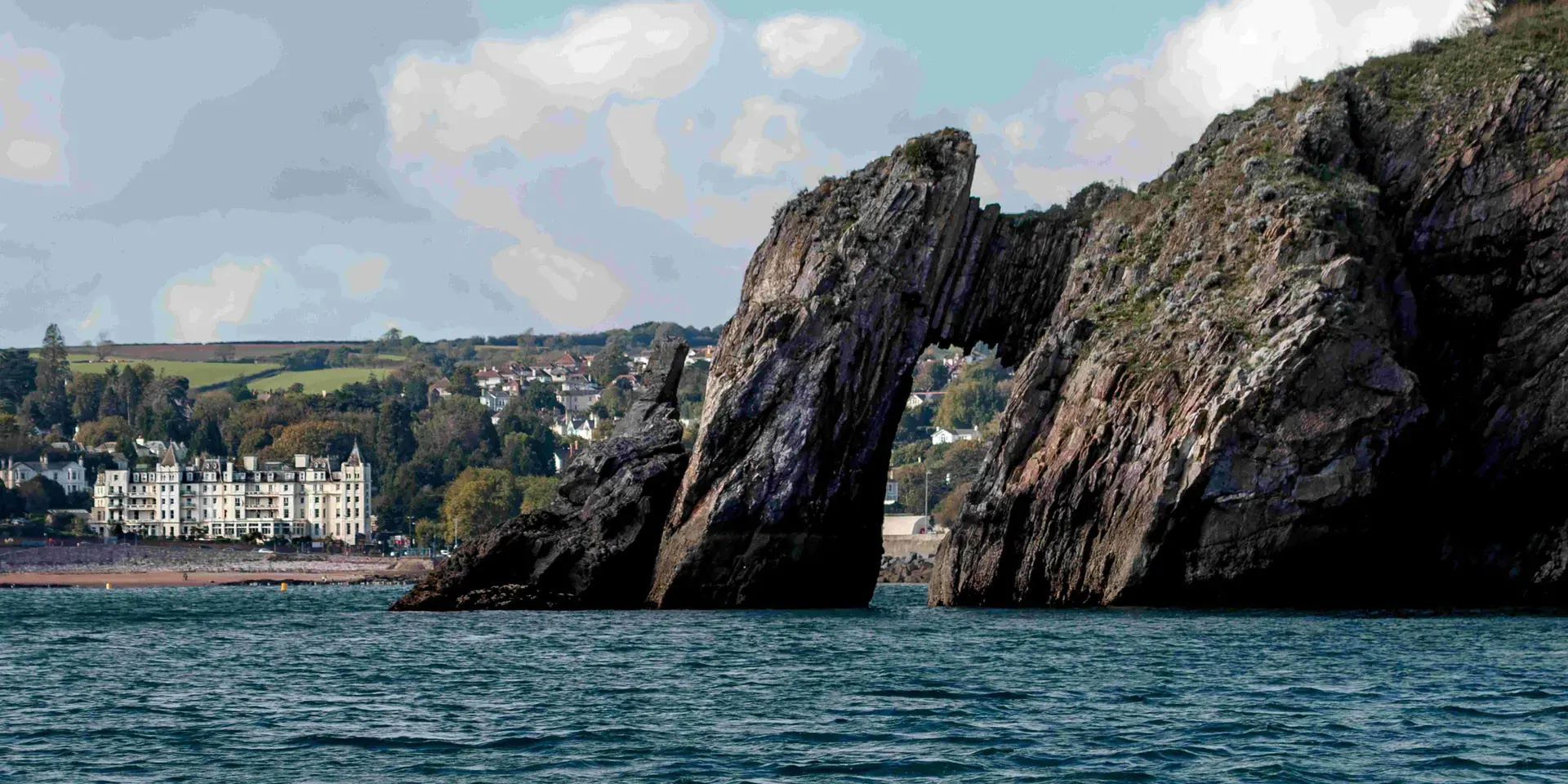 Coastal rock arch rising from the sea with a distant view of a white seaside hotel and green hills under a partly cloudy sky.
