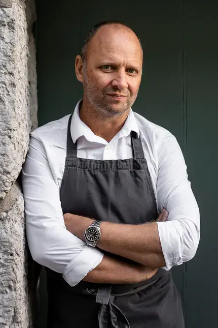 Chef Simon Rogan leaning against a stone wall.