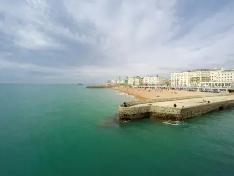 Coastal view with turquoise water, a stone pier, and a sandy beach lined with classic white buildings under a cloudy sky