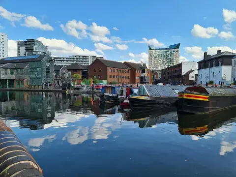 Canal boats docked on calm water, surrounded by historic and modern buildings under a bright blue sky with clouds