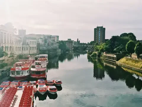 Colorful boats moored on a tranquil river with historic buildings and modern structures under a soft cloudy sky