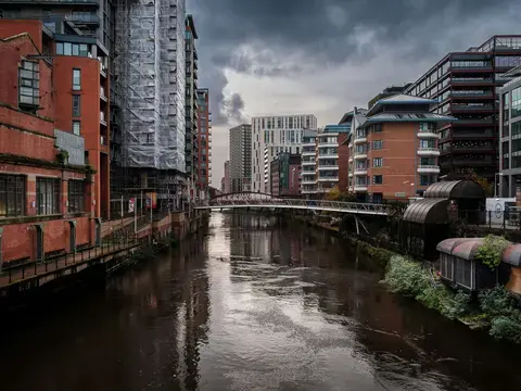 Modern buildings line the river in Manchester’s city center with a pedestrian bridge connecting both sides under a cloudy sky
