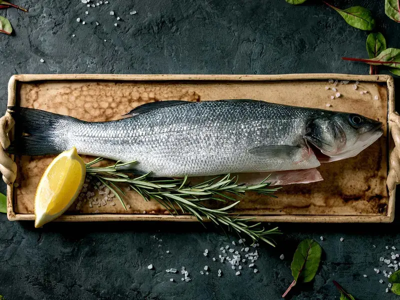 Raw sea bass on ceramic plate with lemon, salt, herbs and leaves.