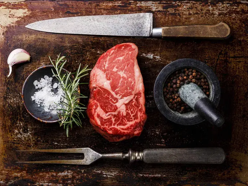 Ribeye steak on a board with seasoning, herbs, and a knife and fork.