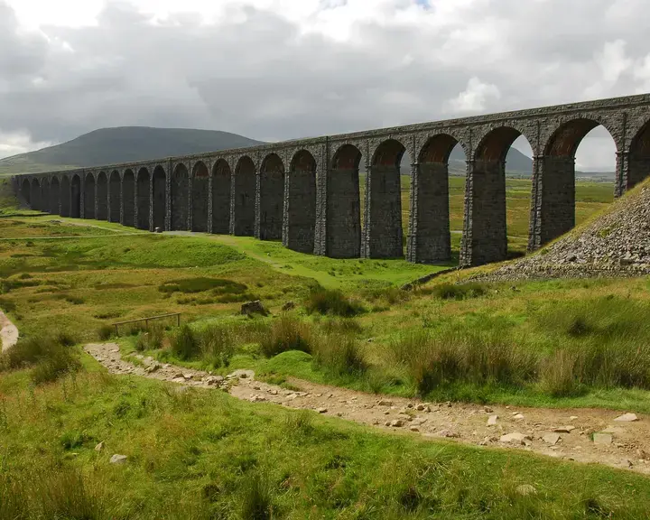 Stone viaduct with arches spans green moorland under cloudy skies, a dirt path winds towards the iconic structure