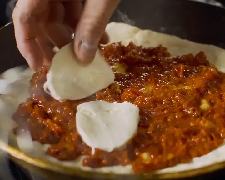 Hand placing slices of mozzarella on pizza dough with tomato sauce in a frying pan
