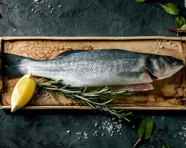 Raw sea bass on ceramic plate with lemon, salt, herbs and leaves.