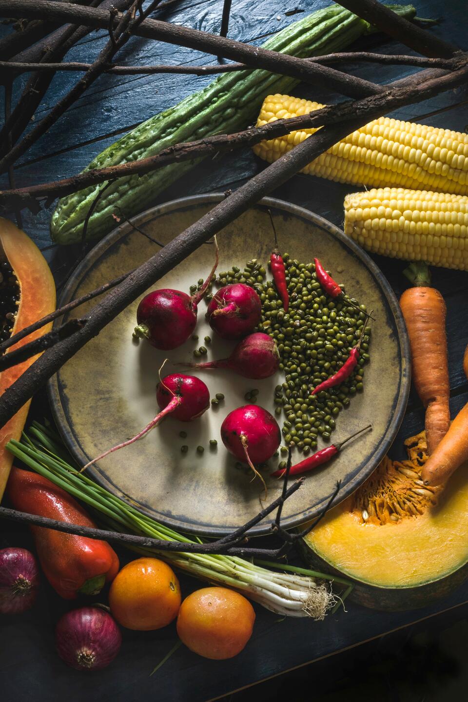 A variety of fresh vegetables including corn, carrots, red onions, cherry tomatoes, and green beans arranged on a dark table